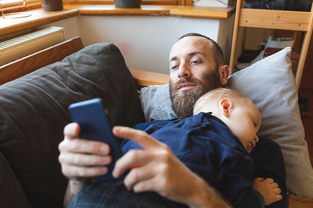 Man checking his phone while his little baby son is sleeping on the sofa