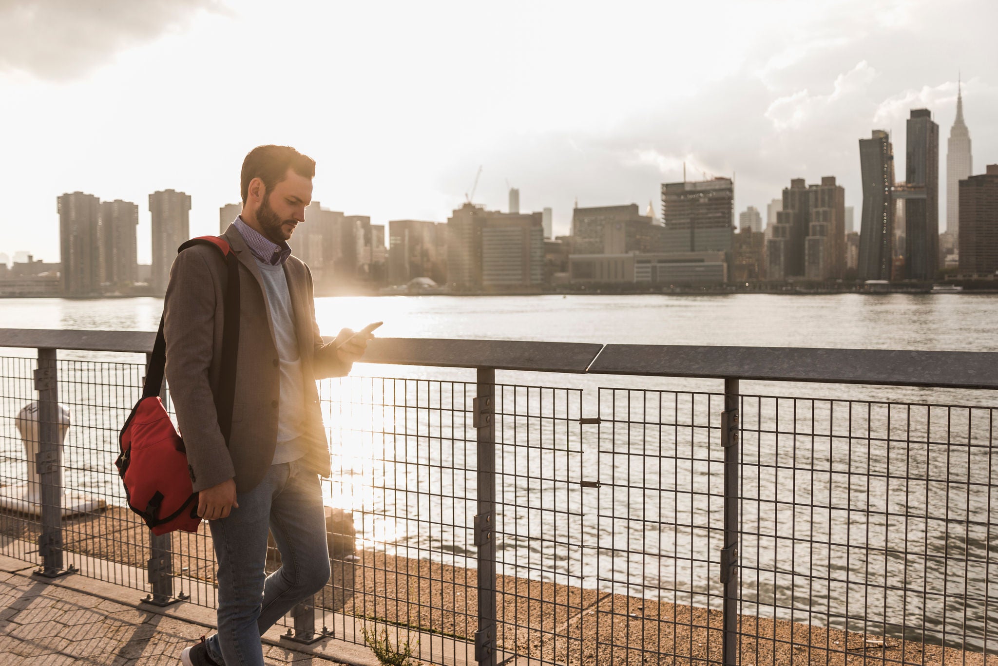 Man walking by waterfront promenade with city skyline at sunset.