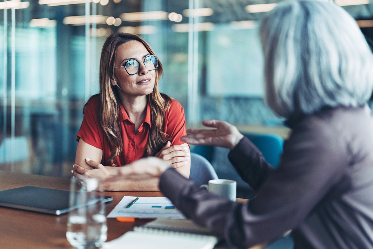 An advisor speaks with a client across a table in a modern office, with documents and a laptop visible, suggesting a professional consultation.