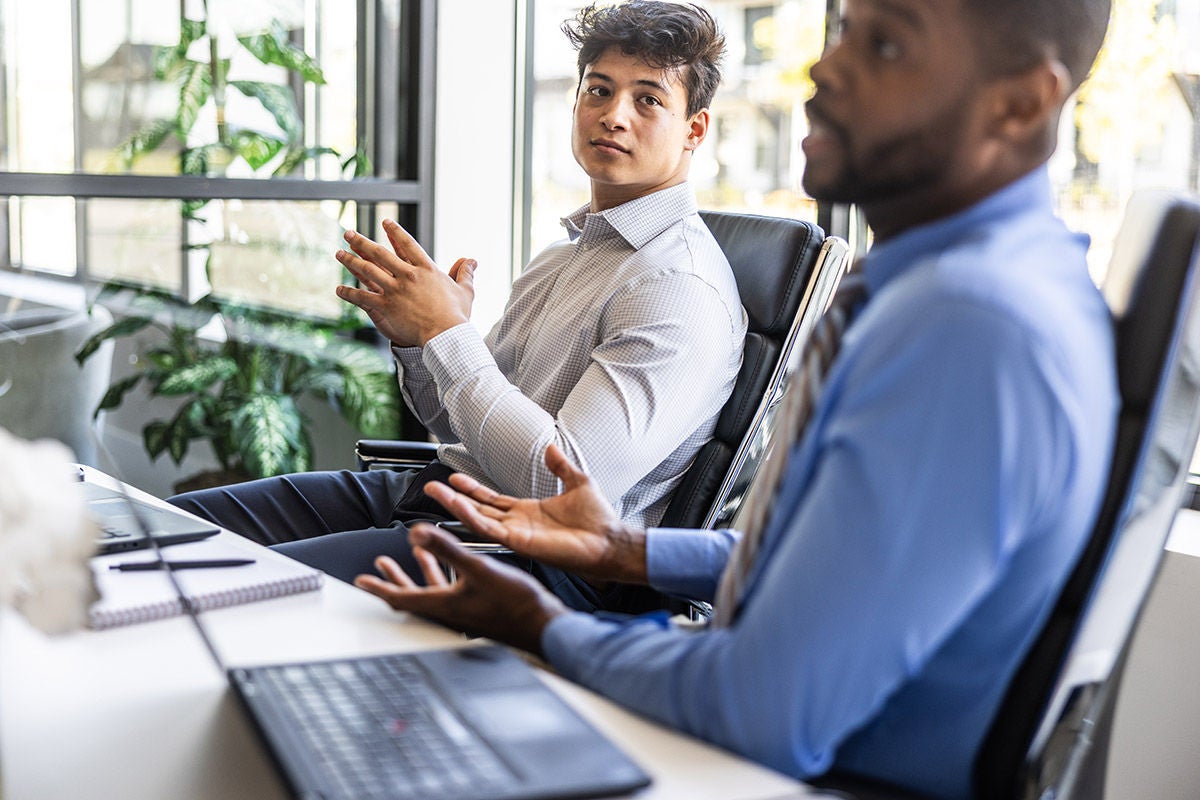 Two professionals sit at a desk in a modern office, gesturing as they discuss ideas, with a laptop, notebook, and large windows letting in natural light.