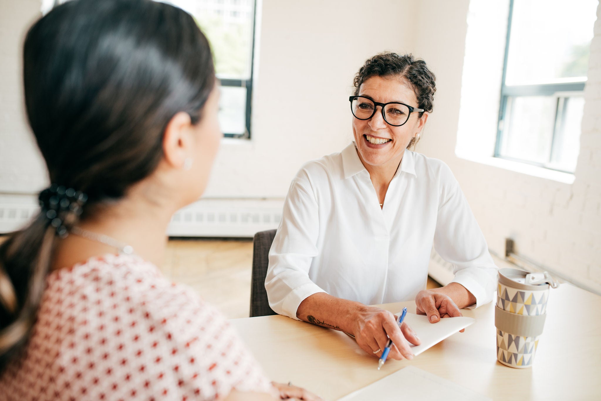 Two people sit across from each other at a table in a bright office, with one person holding a pen and taking notes during a conversation.
