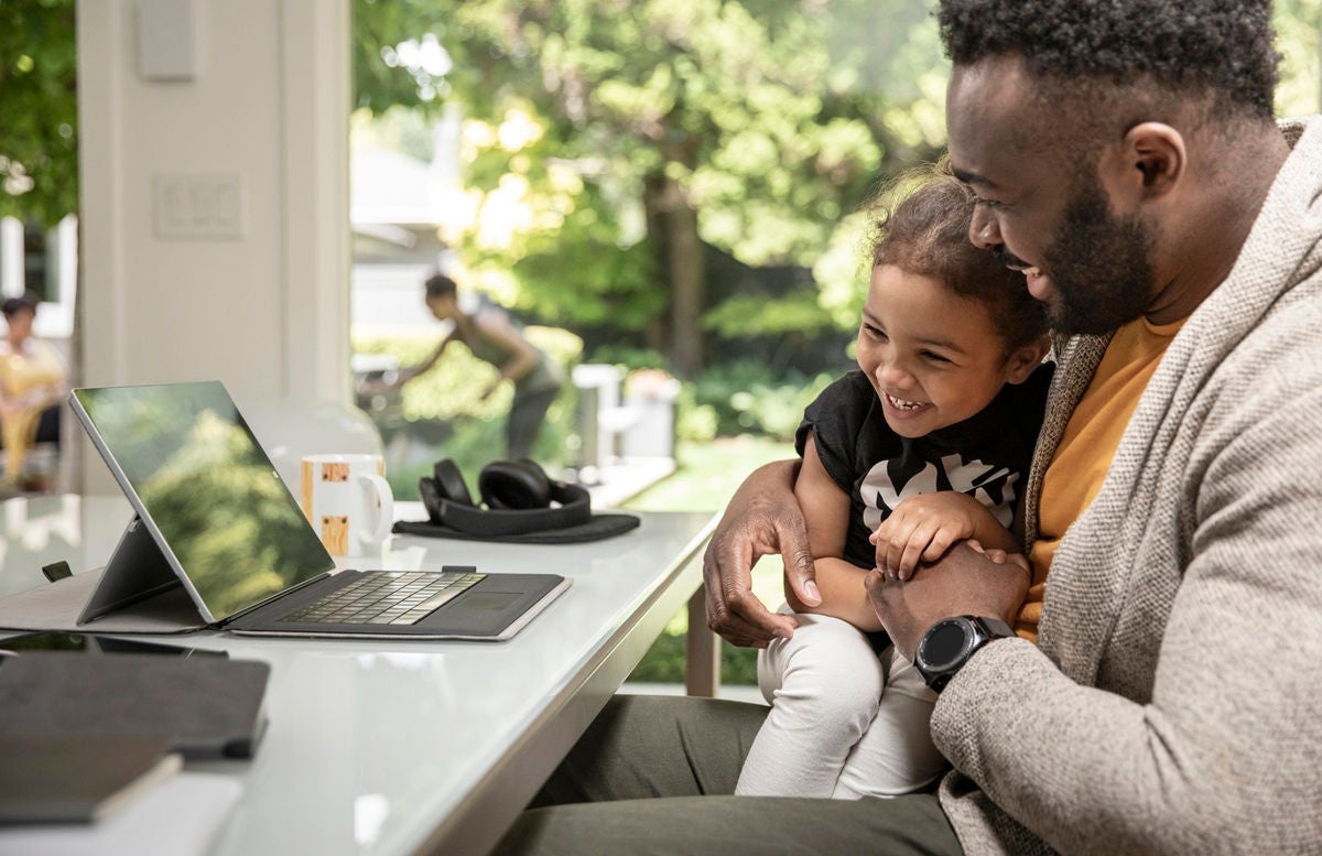 A man is sitting at a desk holding his daughter on his knee and they're both smiling and laughing