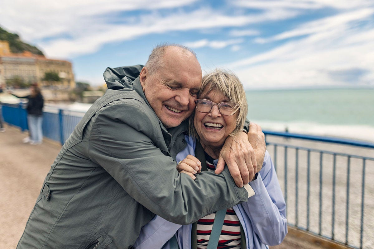 Two people on vacation embracing on a seaside promenade, standing beside a railing with the ocean, shoreline, and buildings visible in the background.