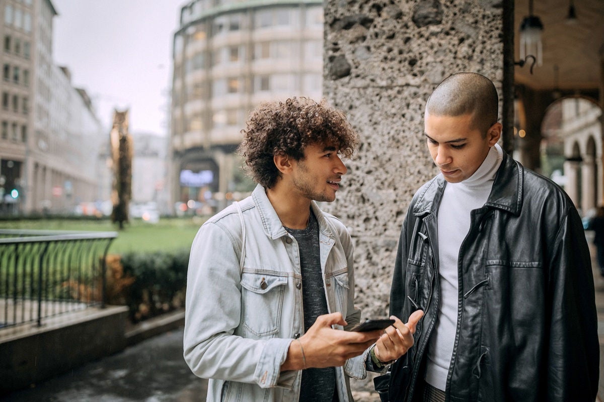 Two friends talking on a city sidewalk while looking at a phone.