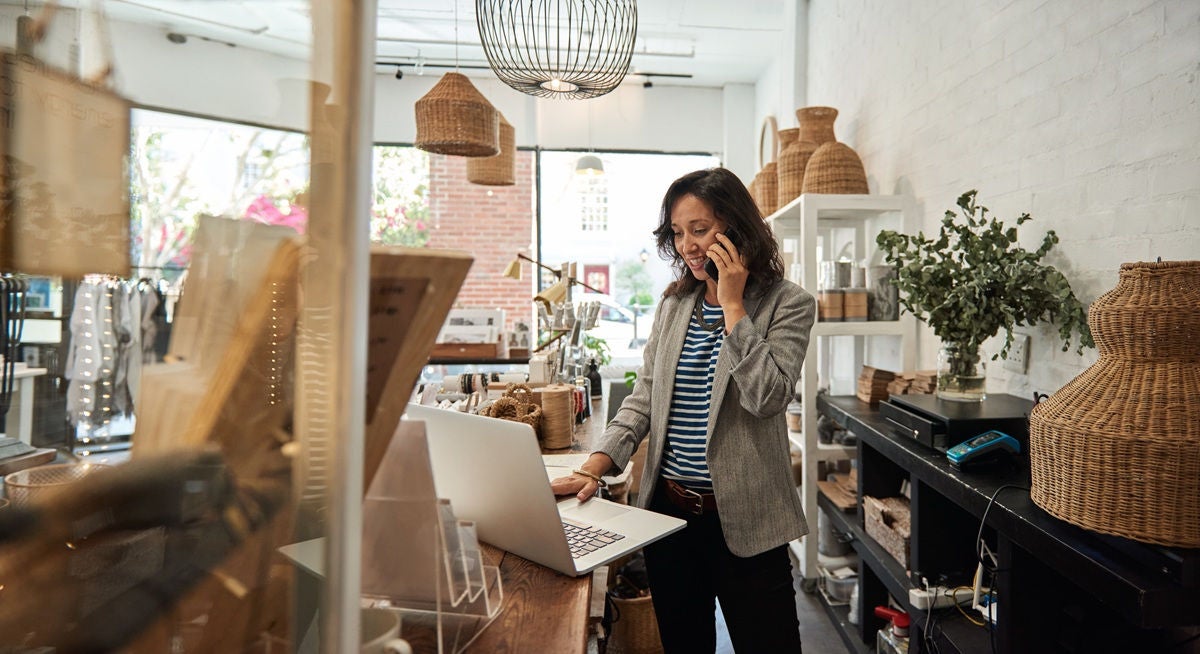 Shop owner standing at a counter in a bright, stylish retail store, using a laptop and phone, with shelves of woven baskets and home décor in the background.