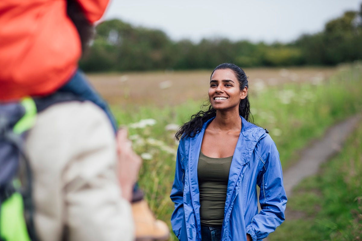 Person in a blue jacket standing on a rural path while another adult with a child is in the foreground, surrounded by fields and greenery.