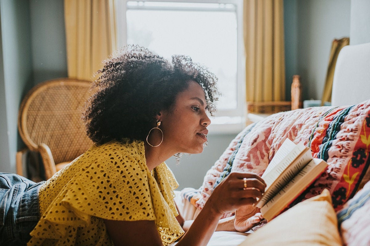A person lies on a bed reading a book in a cozy, sunlit room.