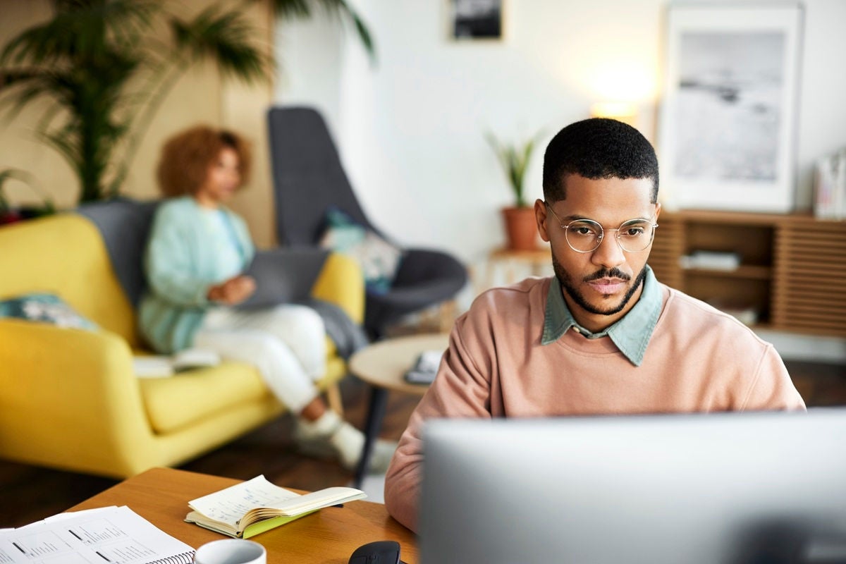 Confident male freelancer using computer at desk. Businessman is working at home office. He is with colleague in apartment.