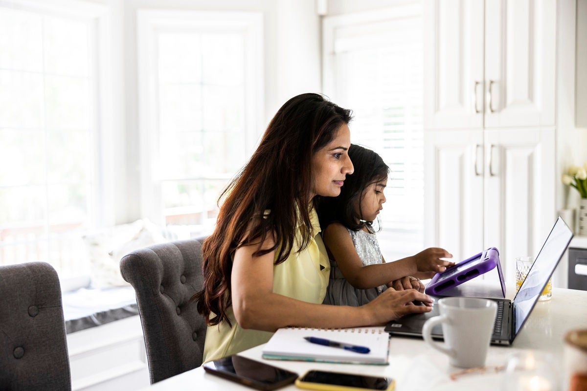 Adult and child sitting at a kitchen counter using a laptop and tablet together, with notebooks, a mug, and a bright kitchen workspace around them.