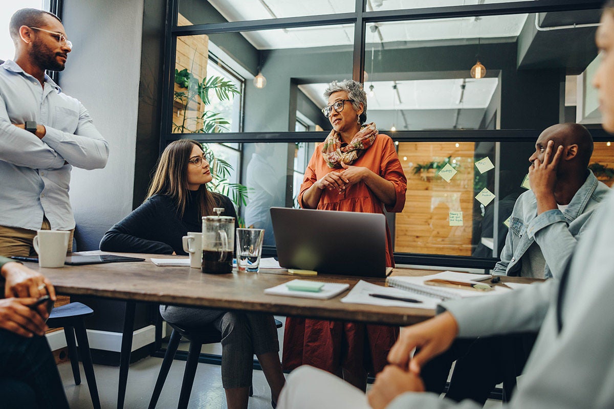 A group of colleagues gather around a table in a modern office, with one person standing and addressing the team during a collaborative meeting