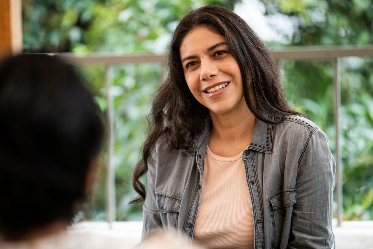 A person with long hair sits outside, listening during a conversation with greenery in the background.
