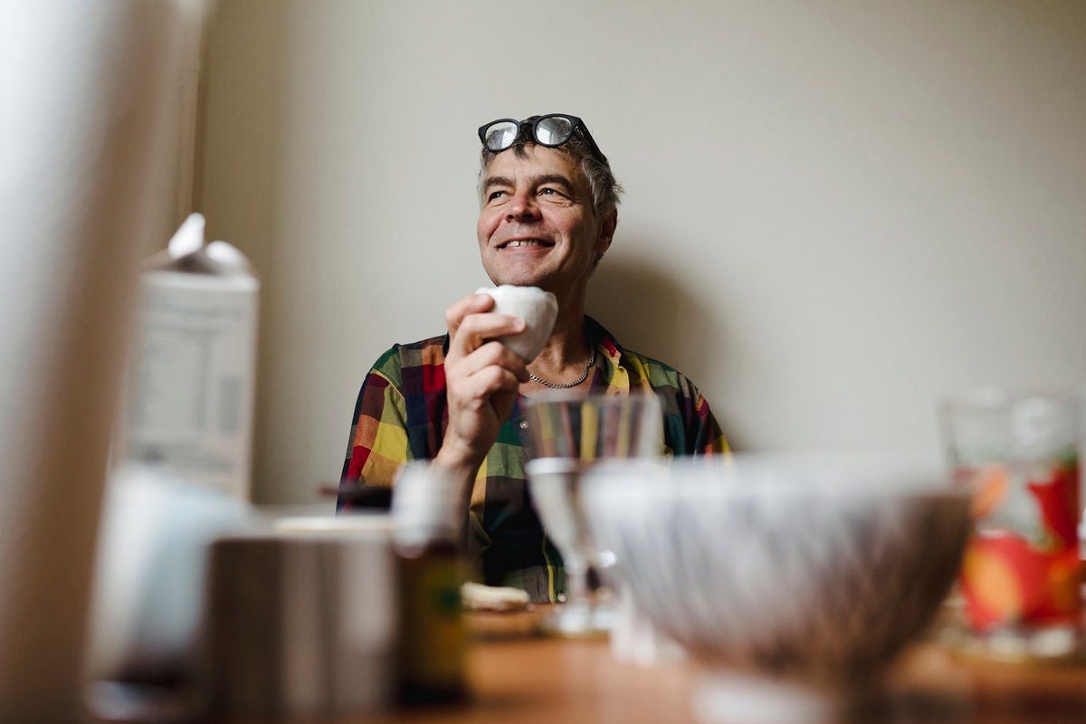 Person sitting at a table holding a cup, surrounded by household items in a casual indoor setting.