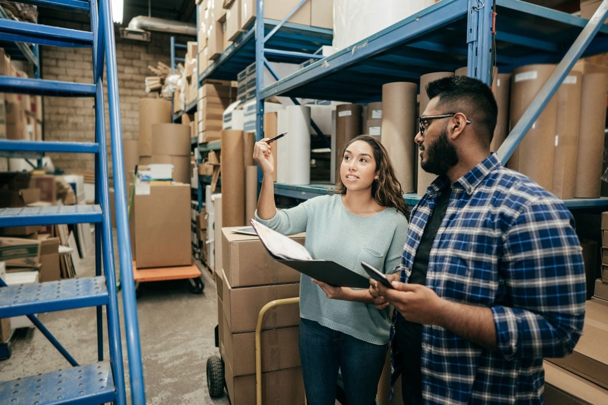 Two coworkers stand in a warehouse aisle surrounded by stacked boxes and shelving, reviewing inventory with a clipboard and pen while discussing items on the shelves.