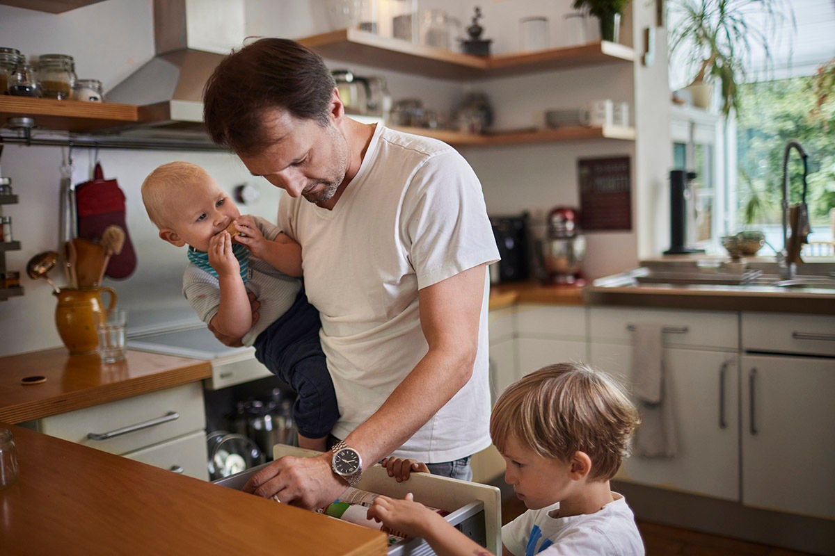 Adult standing in a home kitchen holding a baby while another young child helps open a lower cabinet.