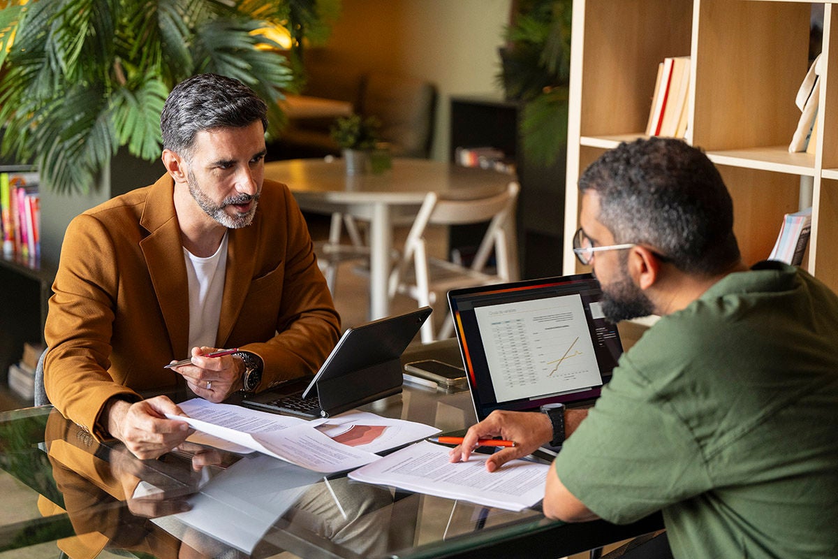 Two people sitting at a table reviewing financial documents and discussing information on a laptop.