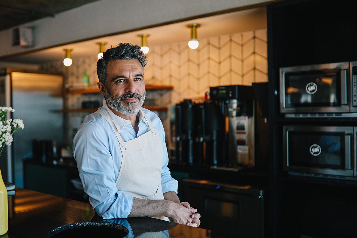 A café or restaurant owner stands behind a counter wearing an apron, with coffee equipment and ovens visible in a warmly lit commercial kitchen.