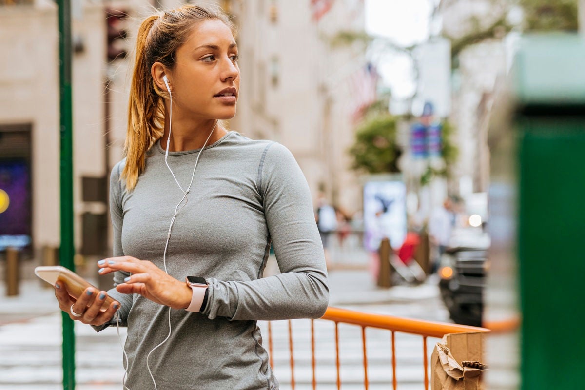 Person standing on a city sidewalk wearing athletic clothing, listening to earphones, and using a smartphone near a street crossing