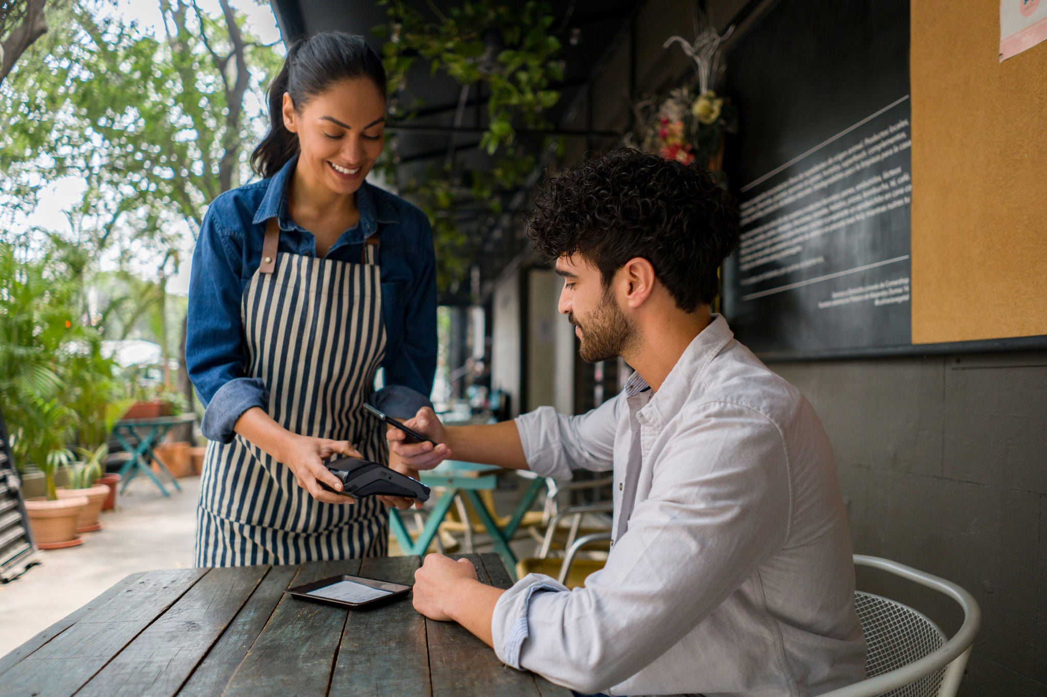 A server wearing an apron hands a payment terminal to a seated customer at an outdoor café table surrounded by plants and casual seating.
