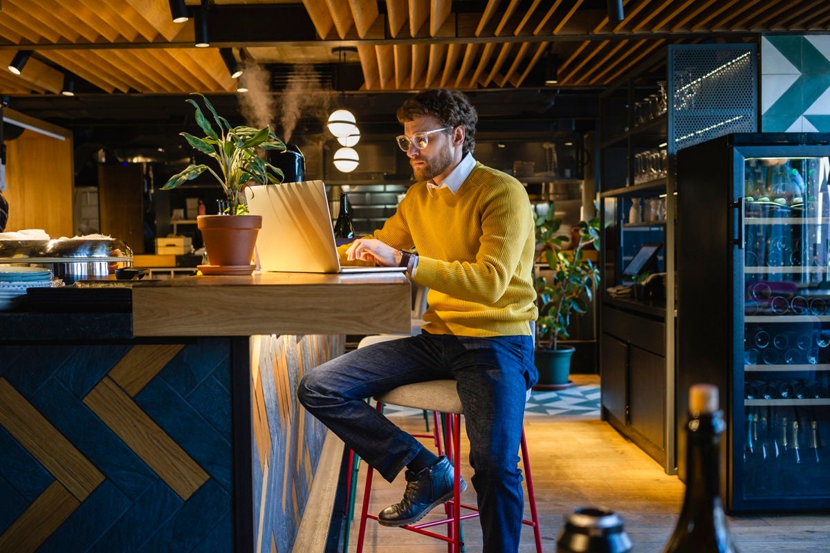Person working on a laptop at a wooden counter in a modern café with warm lighting and plants.