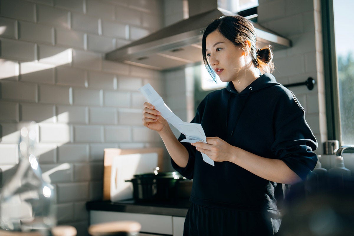 Person standing in a kitchen reading a bil, with a stovetop and tiled backsplash visible in the background.