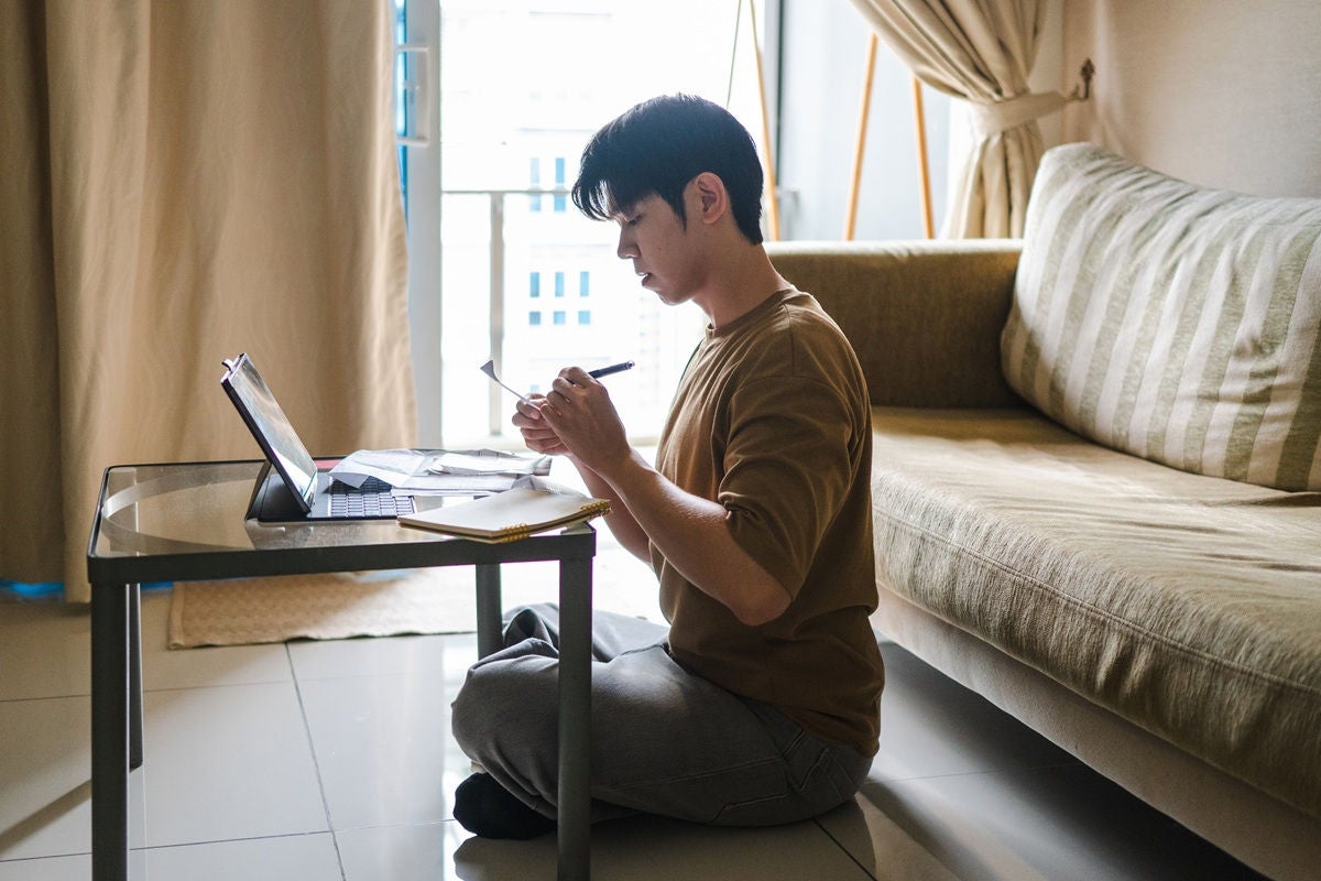 A person kneels on the floor working at a low table with a laptop, papers, and a calculator in a bright living room.