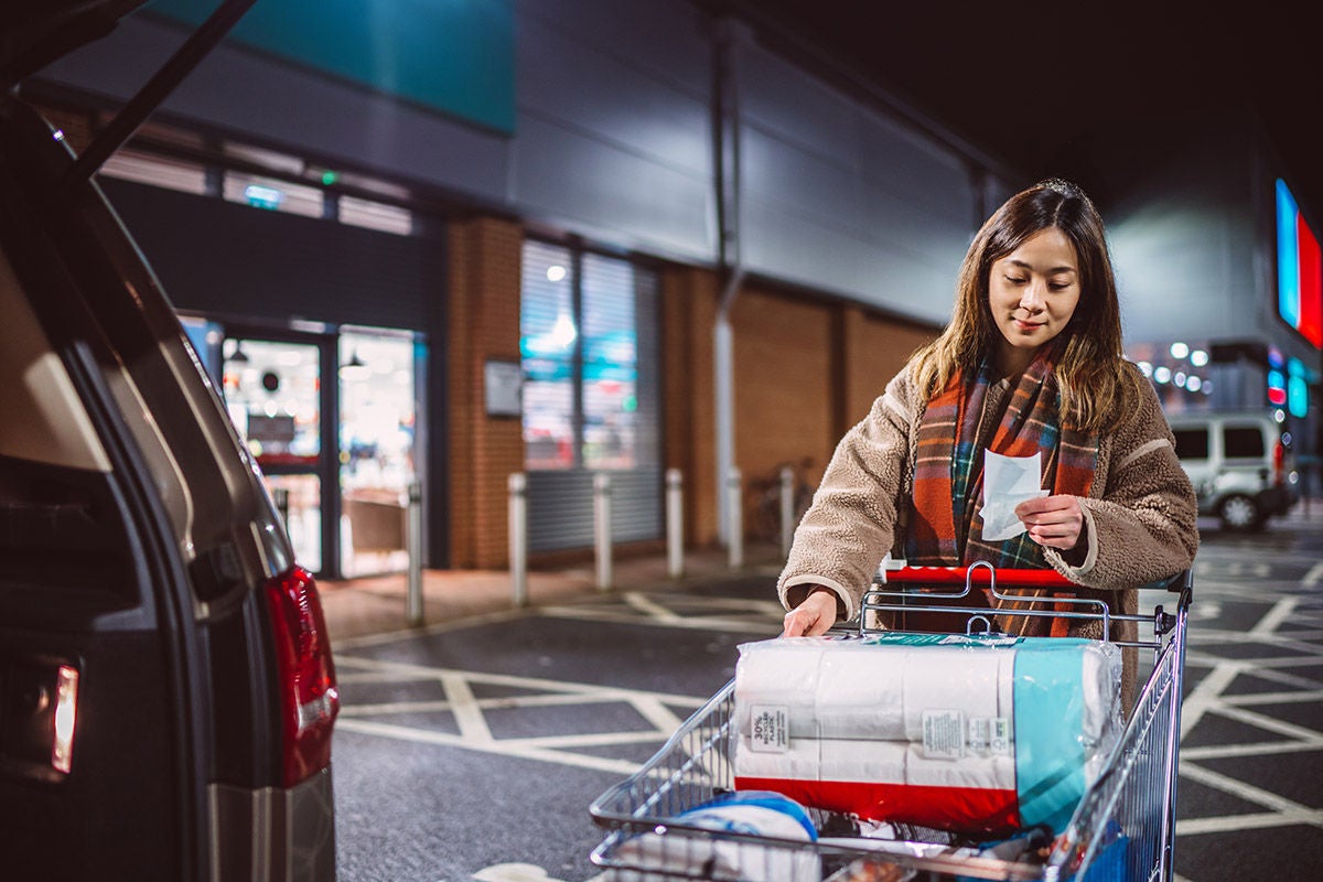 Woman checking her shopping receipt beside a supermarket parking lot at night, loading household essentials into her car. Concept for inflation, family budgeting, sustainability, and mindful spending.