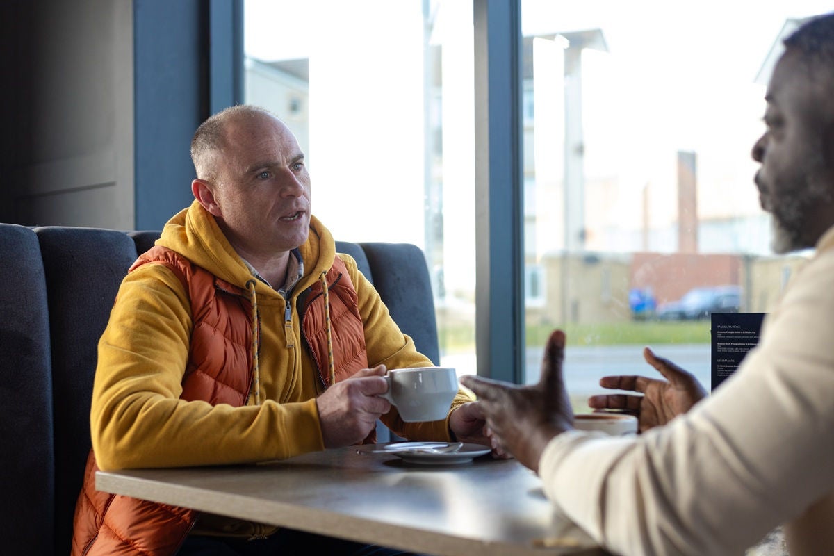 Medium over-the-shoulder shot of two men drinking a cup of coffee while having a talk. They are sitting at a table together in a cafe. The man on the right is a Polio survivor and lives with disabilities. 

Videos are available similar to this scenario.
