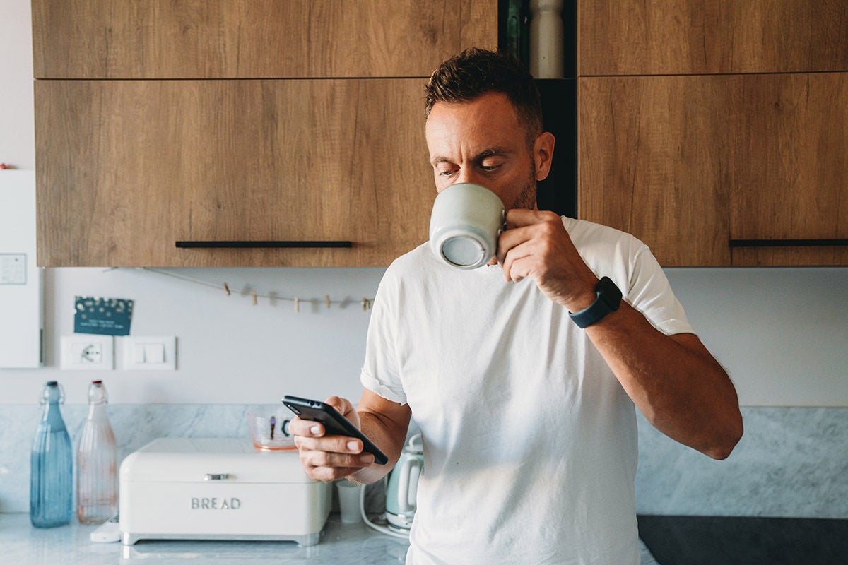 Man standing in a modern kitchen, holding a coffee mug and checking his smartphone, with wooden cabinets and countertop appliances in the background