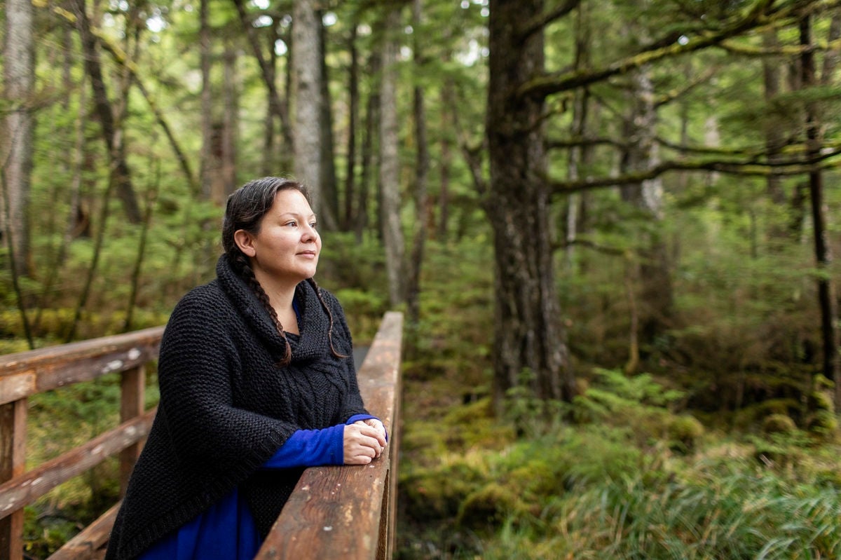 Person standing on a wooden bridge in a lush forest, resting their hands on the railing and looking out over the surrounding greenery.