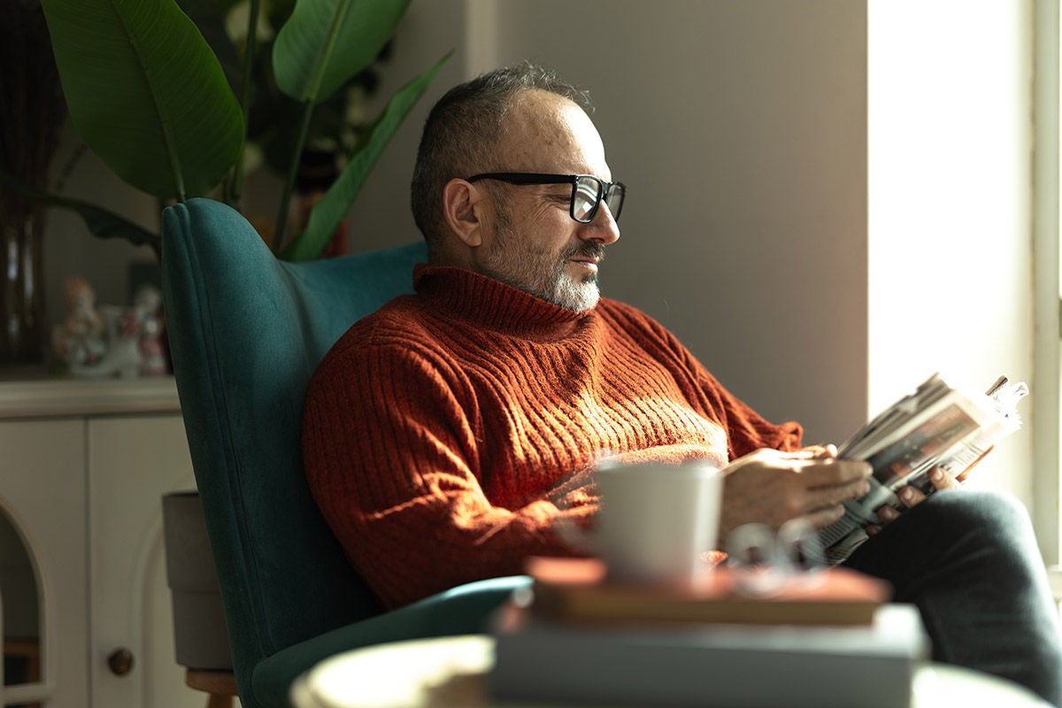 Person sitting in a cozy chair by a bright window, reading a magazine with a coffee nearby.