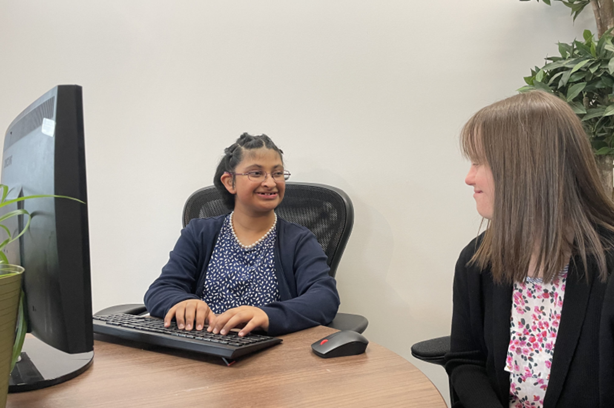 Two people seated at a desk in an office, one typing at a desktop computer while the other listens, with a mouse, keyboard, and plants visible in the workspace.