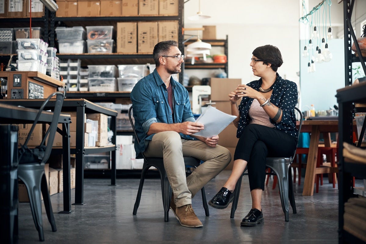 Small business and financial advisor sit in a workshop-style office with shelves of supplies behind them, holding documents and having a focused discussion about financial plans.