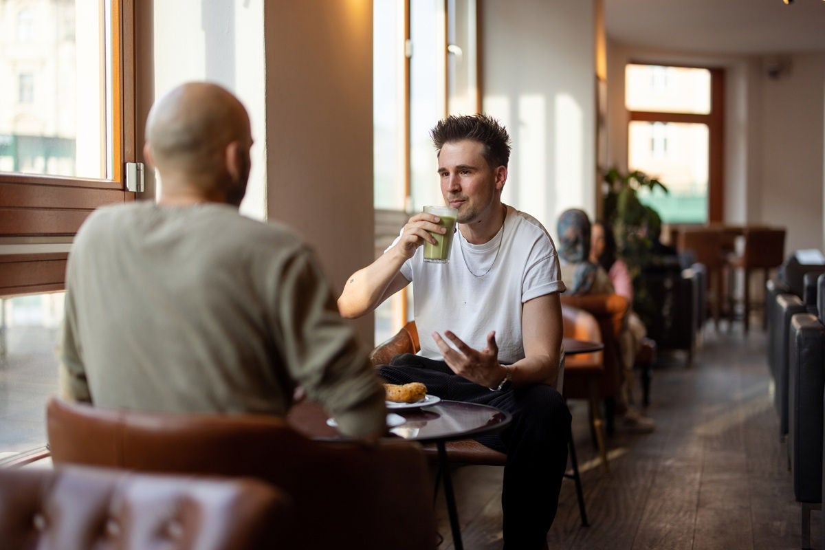 Two people sit at a café table by a window, talking while one drinks from a glass, with other patrons seated along the warmly lit interior behind them.