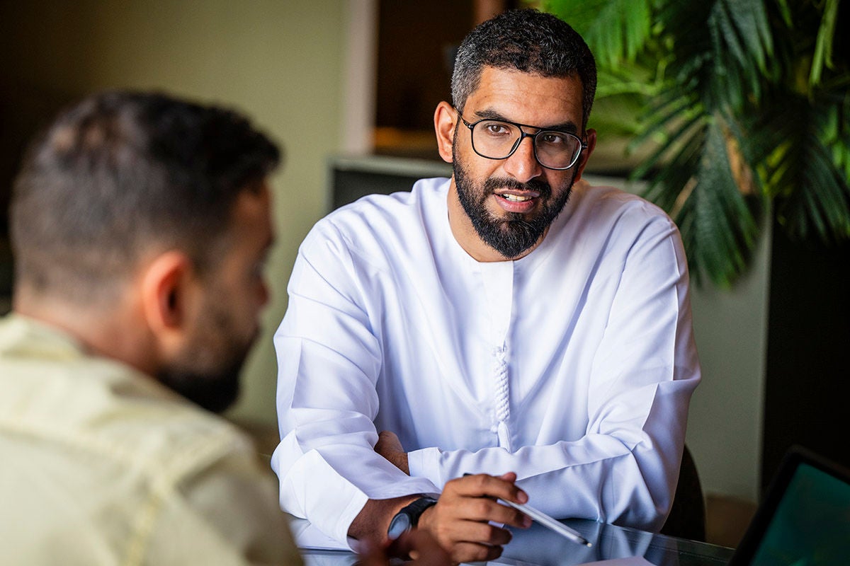 Two people seated at a table in an office setting, engaged in a conversation, with one person holding a pen while listening attentively.