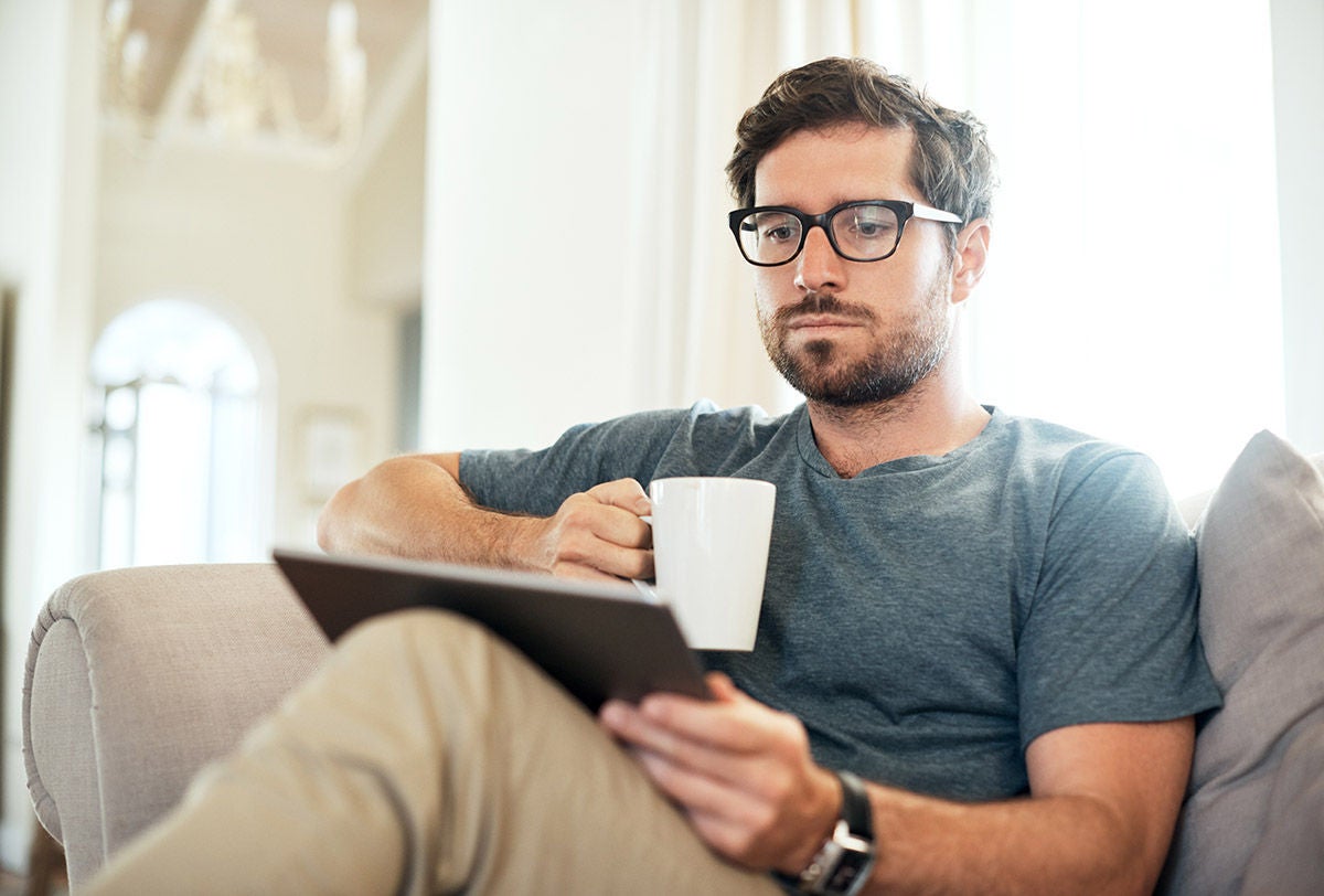 Person sitting on a sofa at home, holding a mug and using a tablet in a bright, comfortable living room.