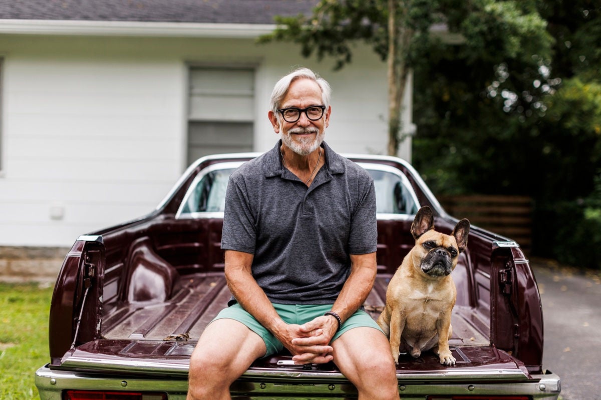 A person sits on the tailgate of a classic truck with a small dog seated beside them in a yard.