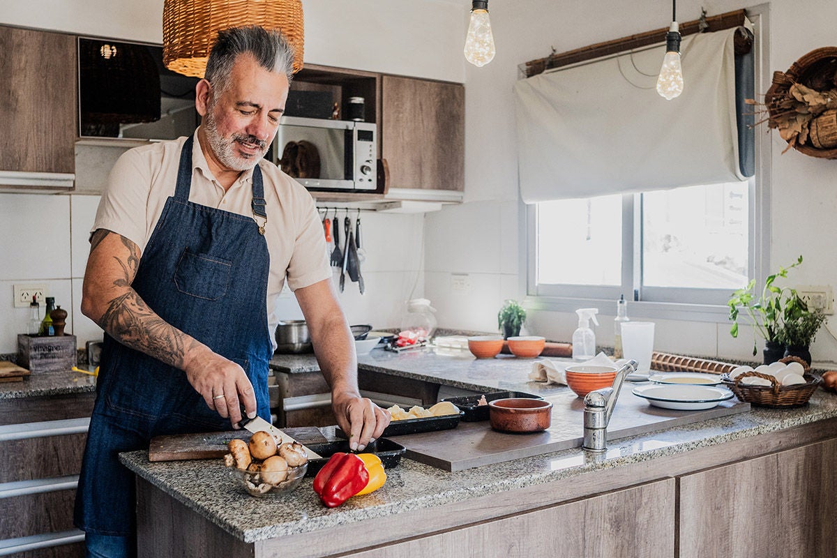 A person in an apron slices mushrooms and a red bell pepper on a kitchen island in a bright, modern kitchen.