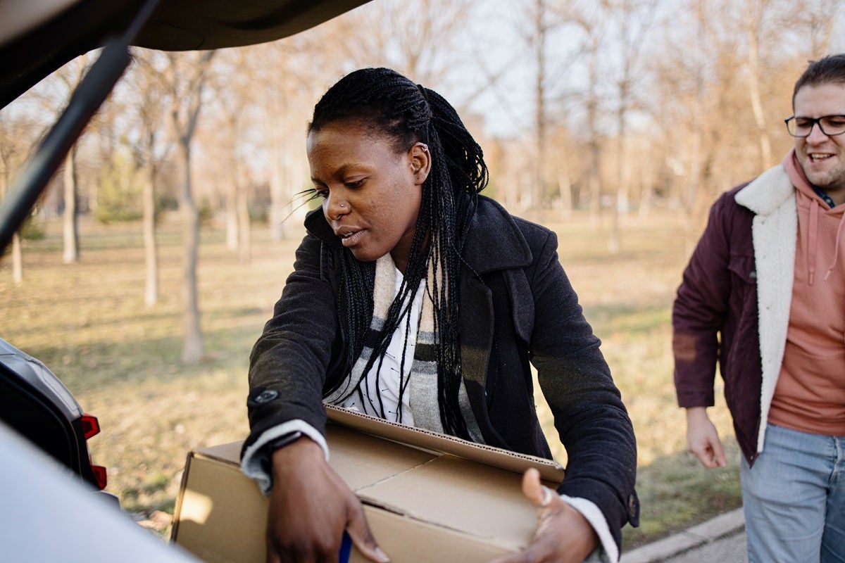 Person lifting a cardboard box from an open car trunk while another person stands nearby in a park setting.