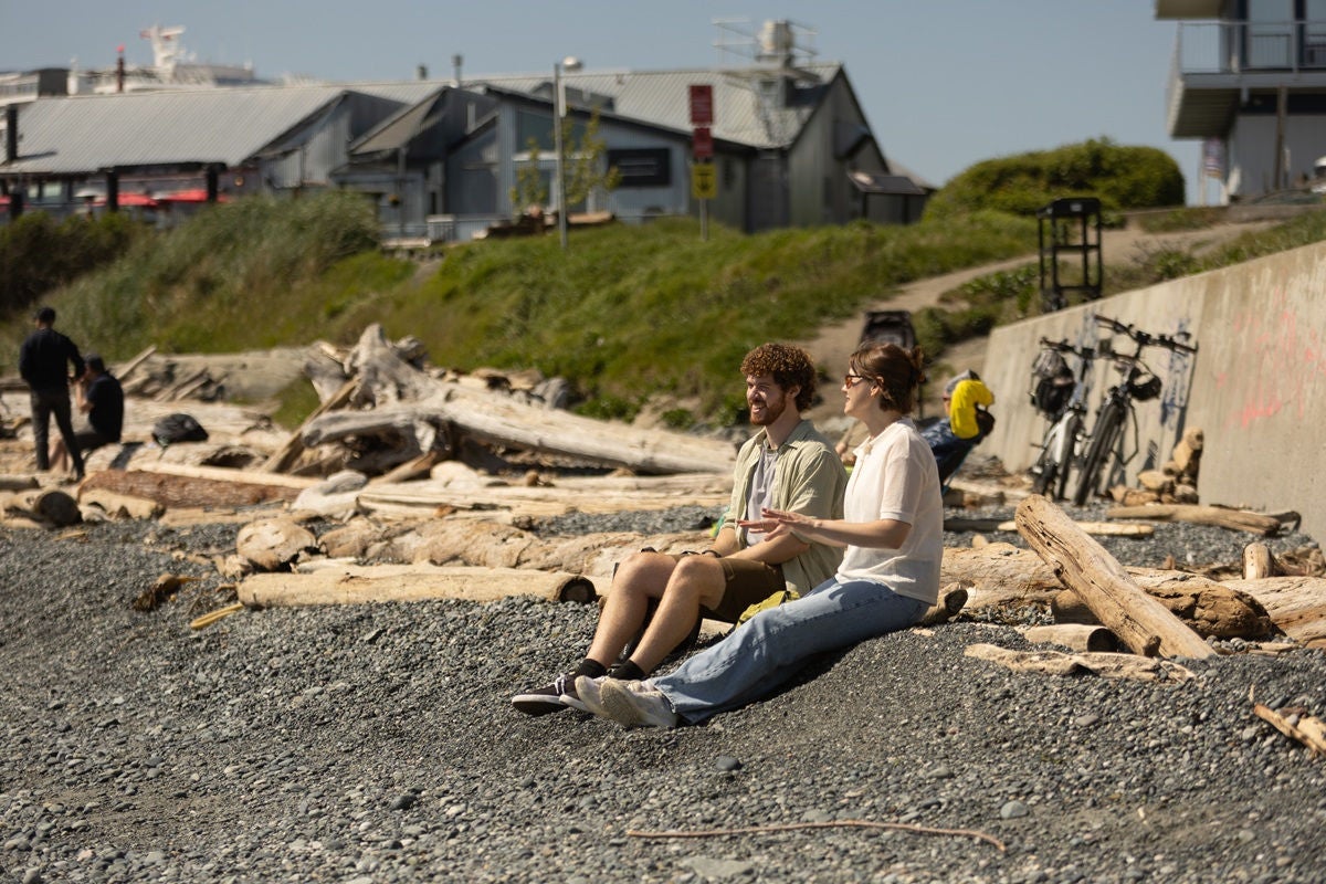 Two people sitting on a rocky beach among driftwood, talking together while looking out toward nearby buildings and the shoreline.