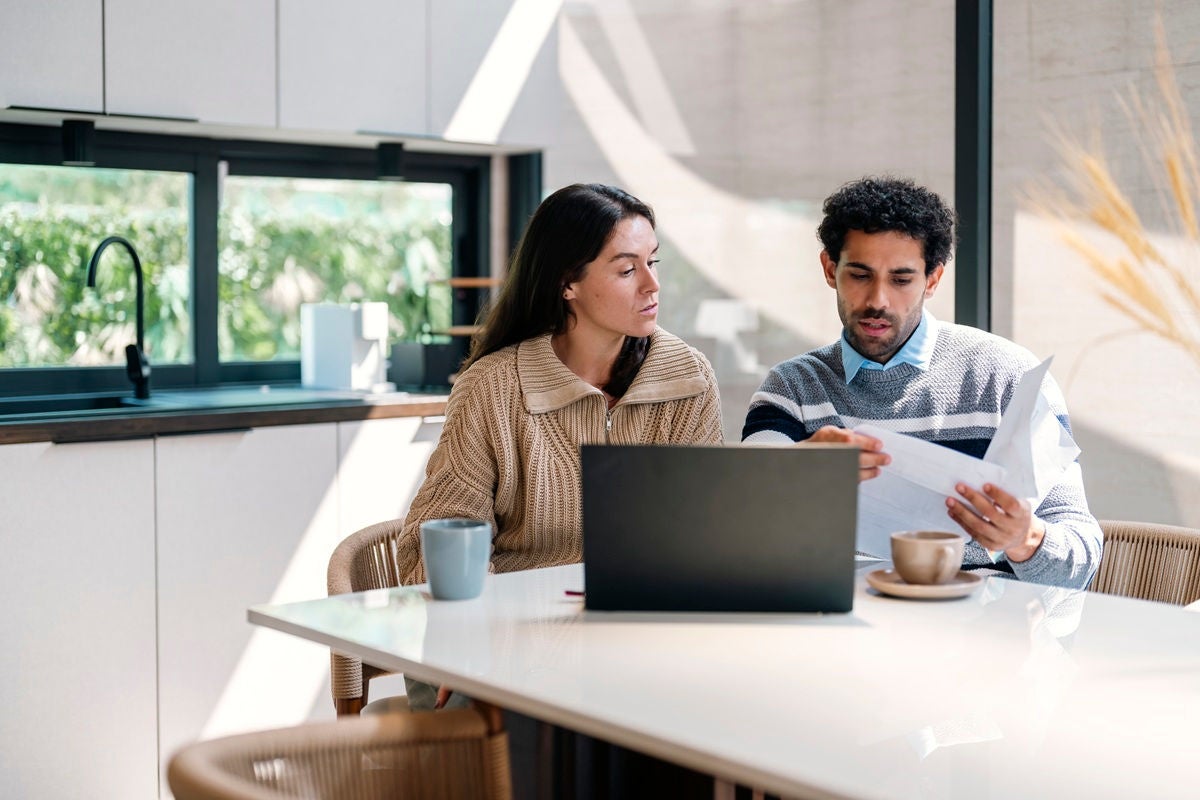 A couple sits at a kitchen table reviewing financial documents together with a laptop open.