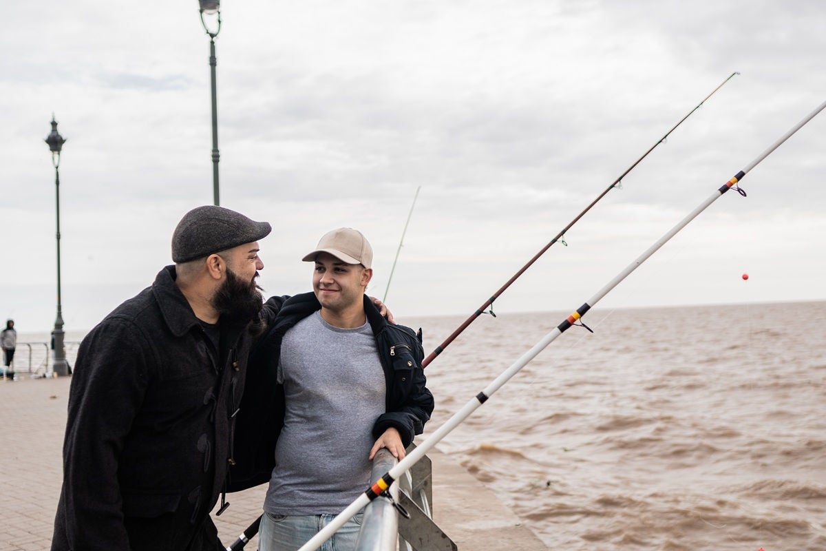 Two people stand together on a waterfront pier, holding fishing rods and looking out over the water on an overcast day.
