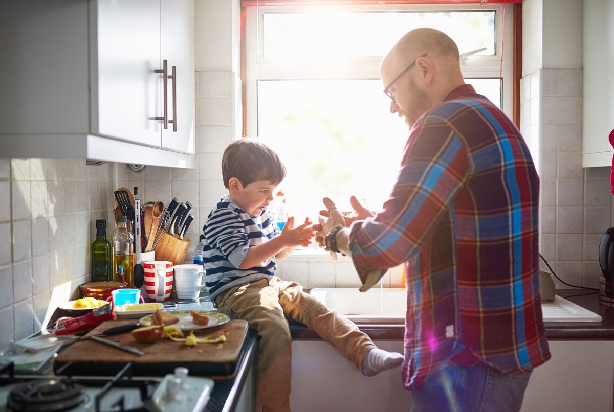 A person helps a young child wash their hands while the child sits on the kitchen counter in a sunlit room.