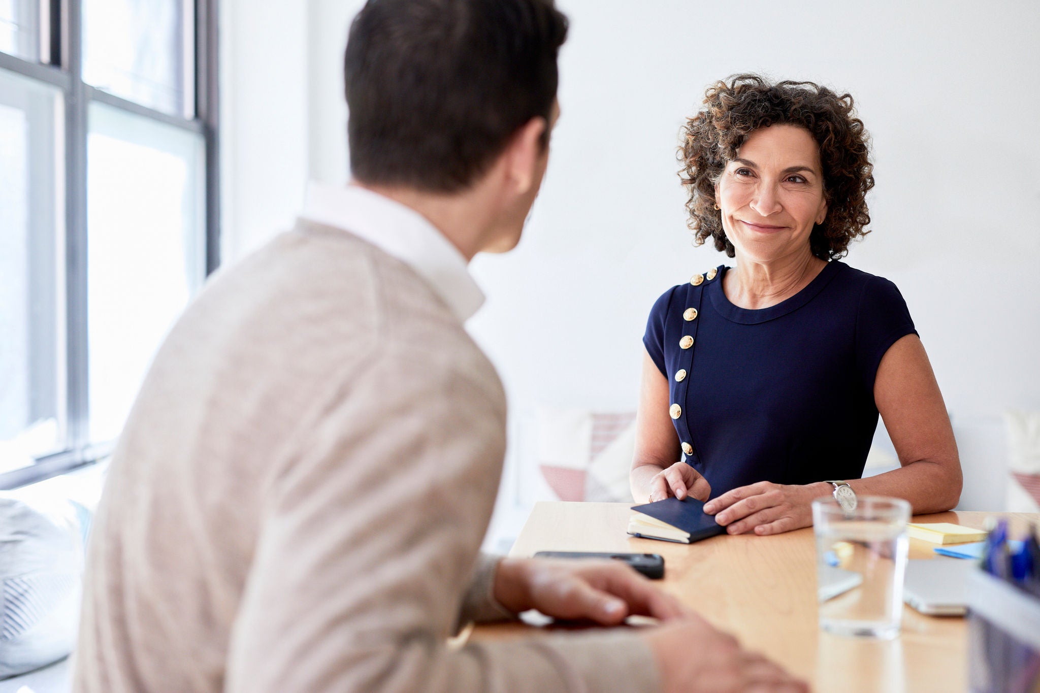 Two professionals sit across from each other at a desk in a bright office, engaged in a focused conversation, with a notebook, phone, and glass of water on the table near a window.
