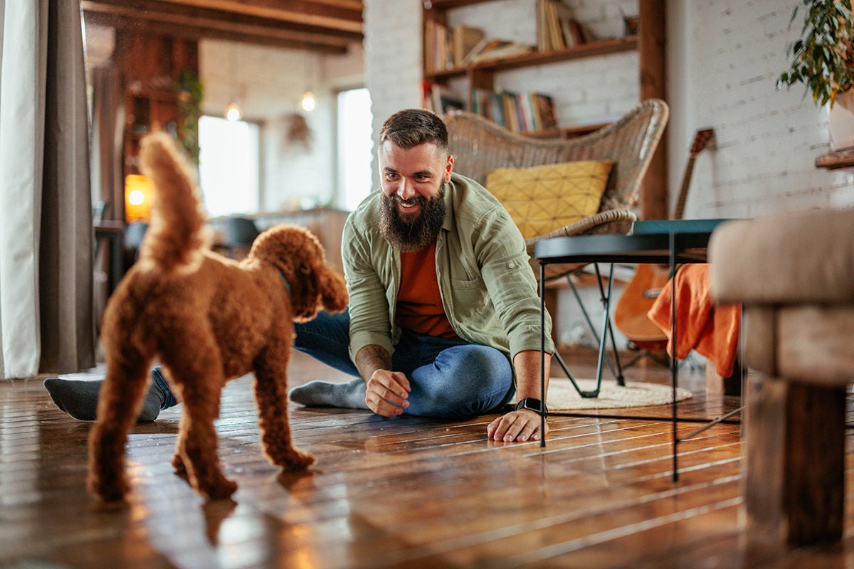 A joyful Caucasian man is at home with his dog playing on the floor in their living room.