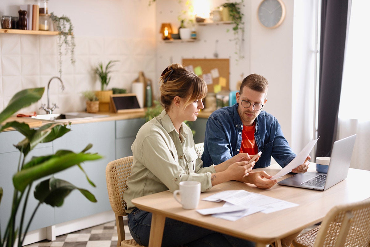 Two people sitting at a kitchen table reviewing documents beside a laptop in a bright home workspace.