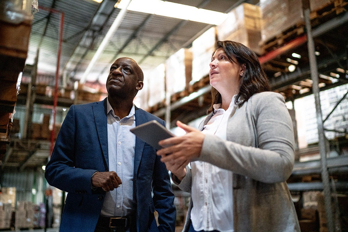 Workers discussing inventory in a warehouse aisle surrounded by stacked pallets.