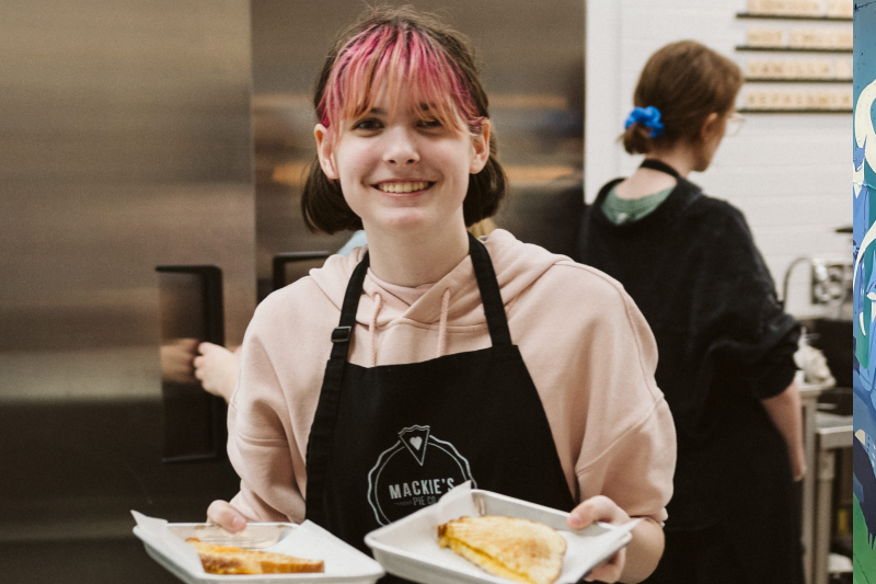 Food service worker wearing an apron and holding two plates of sandwiches in a busy kitchen, with another staff member preparing food in the background.