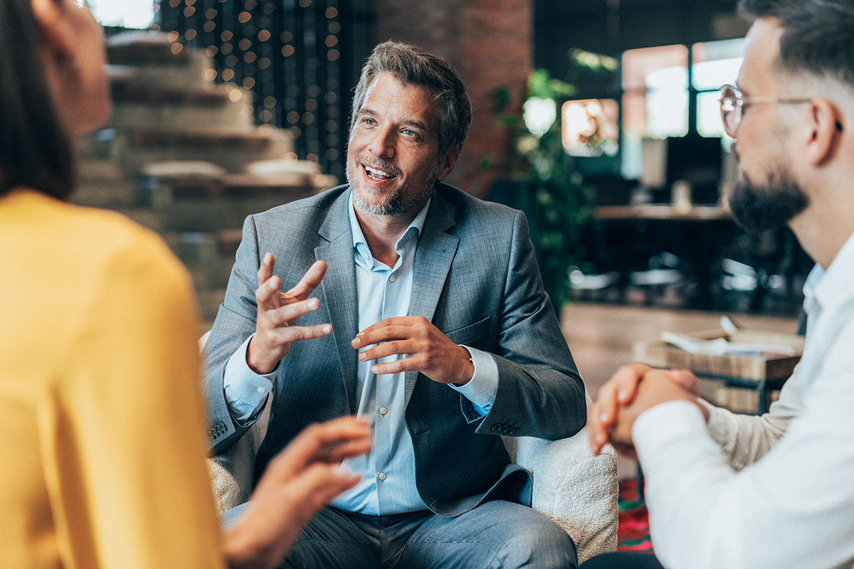 Financial advisor and clients sit in a modern lounge-style office, engaged in an animated discussion, with one person gesturing while the others listen attentively amid warm lighting and contemporary décor.