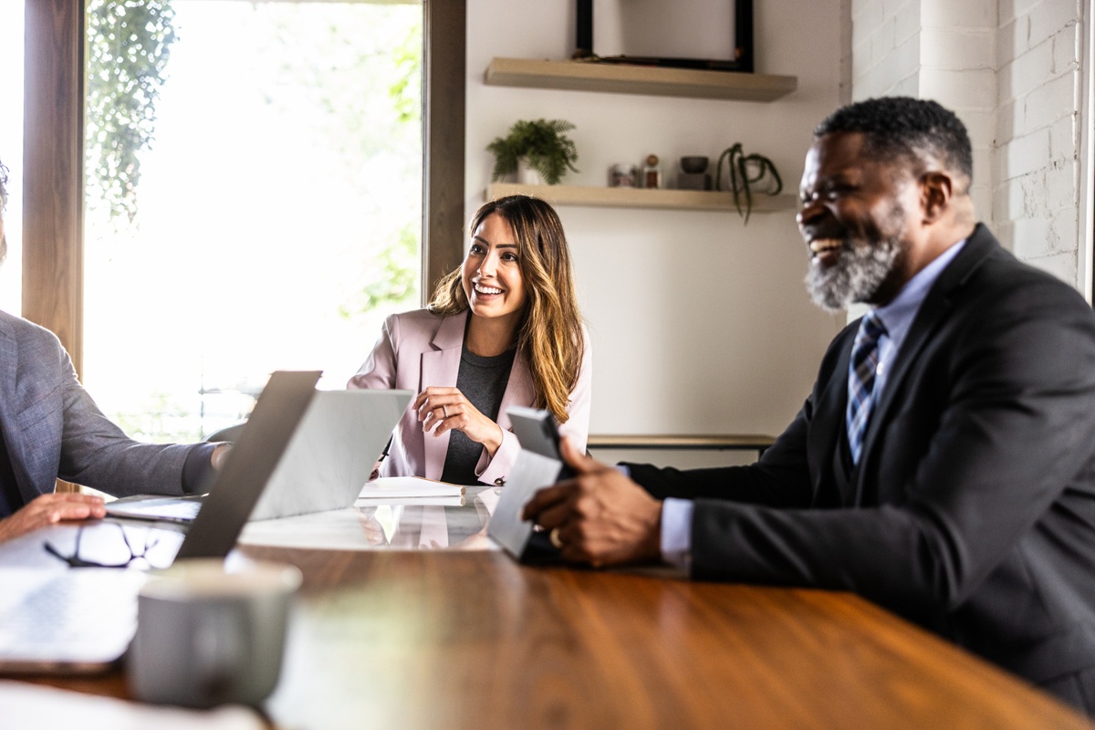 Three professionals sit around a wooden table in a bright office, discussing documents while using a laptop and tablet during a business meeting.