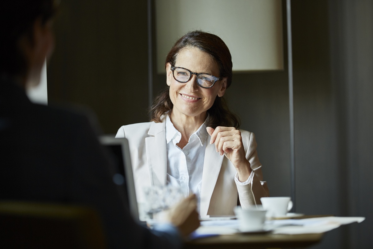 Professional meeting at a table, with a woman in a light blazer speaking across from another person, papers and coffee cups on the table.